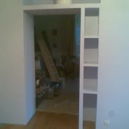 Wnęka w ścianie z półkami, widoczny fragment podłogi i wnętrze pokoju z drabiną i narzędziami, ślady po remoncie.